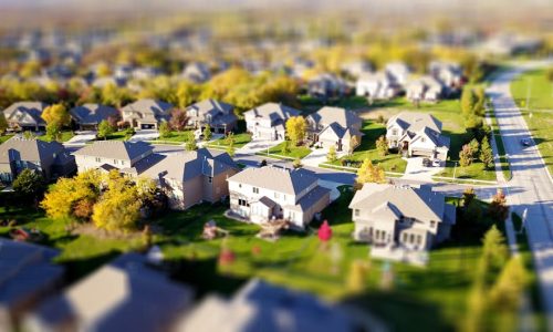 Tilt-shift aerial photo of a suburban neighborhood with green lawns and roads.